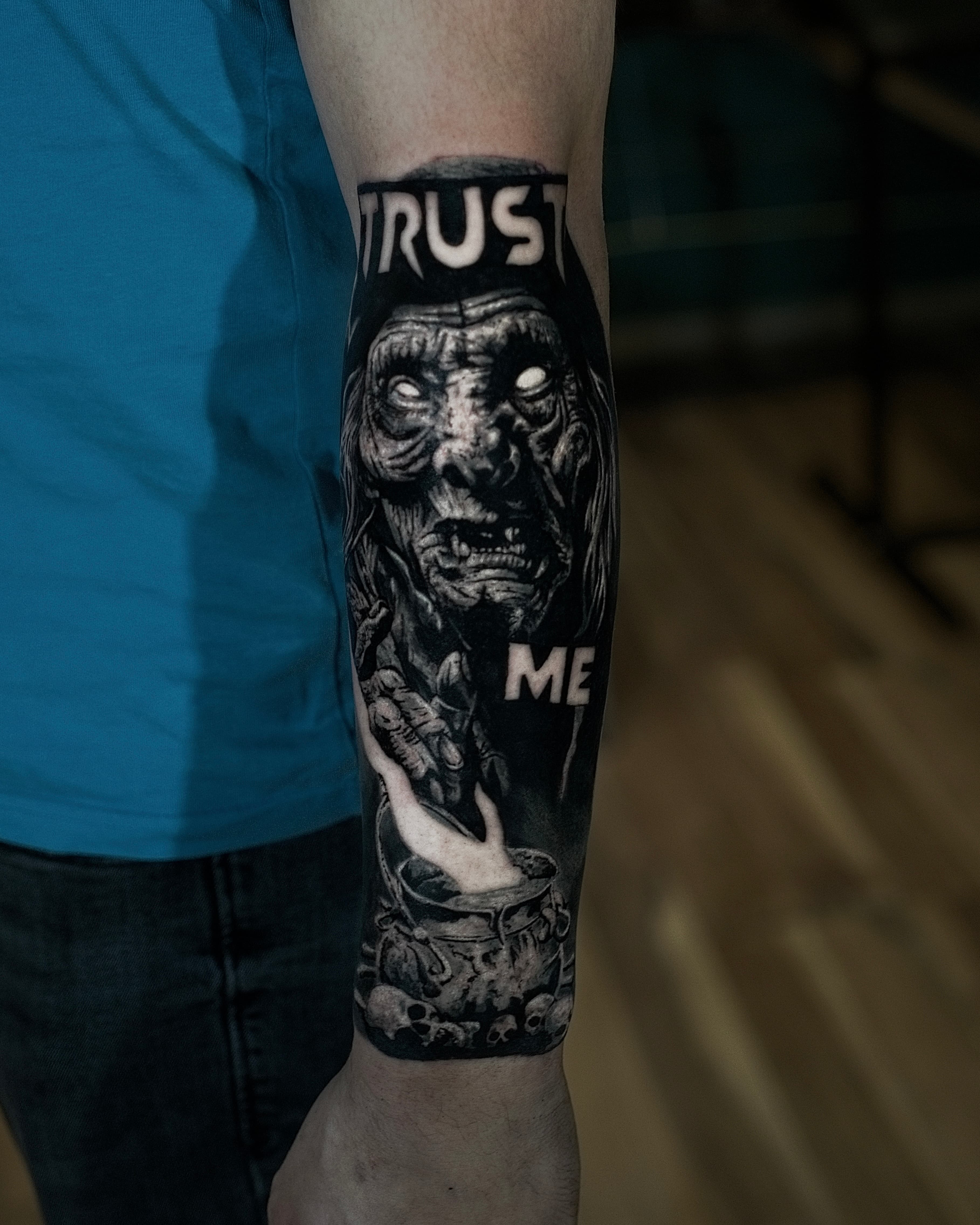 Czarno-szary tatuaż starej wiedźmy nad kotłem z czaszkami i napisem Trust Me
