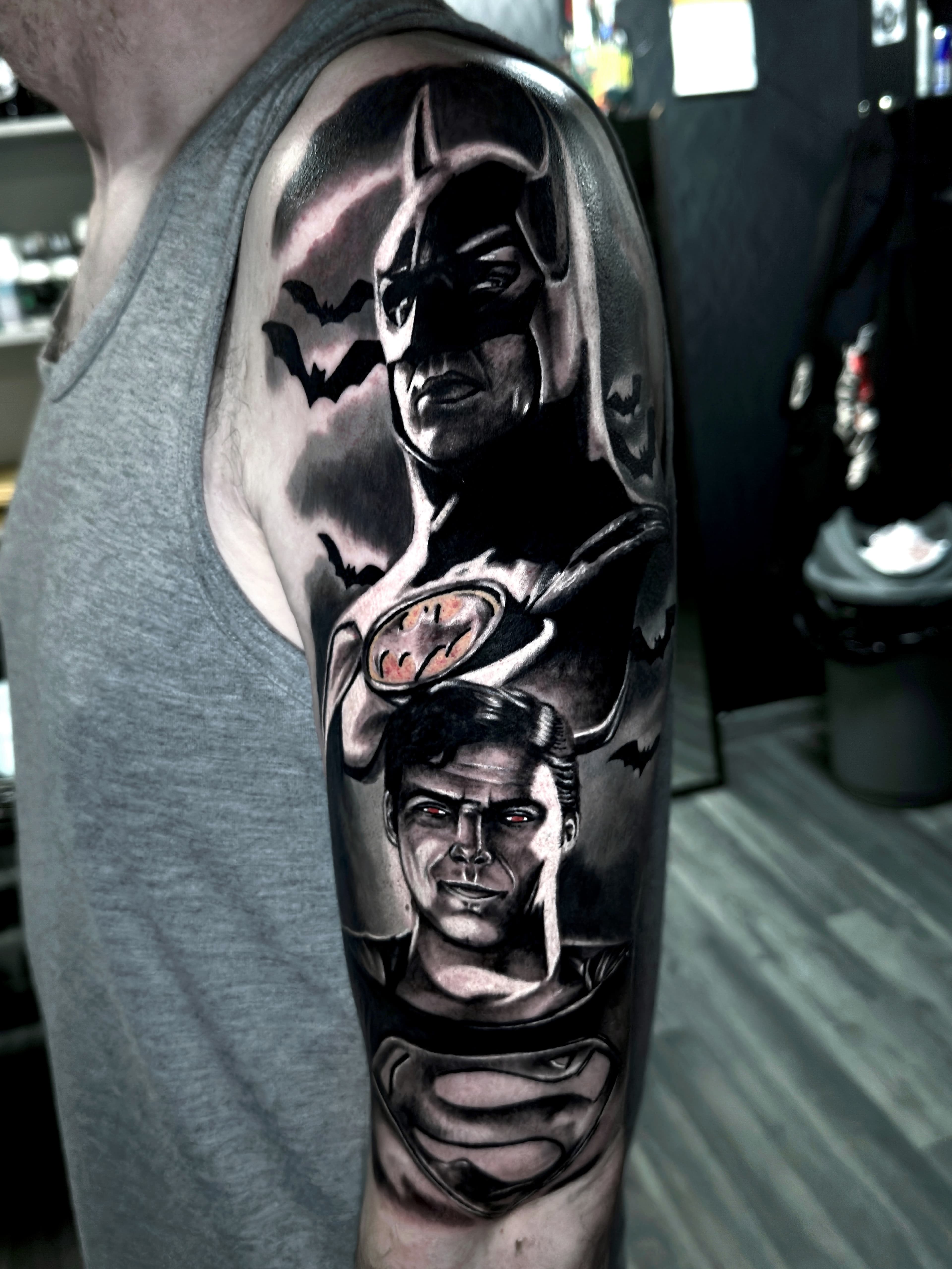 Realistyczny tatuaz Batmana i Supermana w odcieniach szarosci na ramieniu z nietoperzami