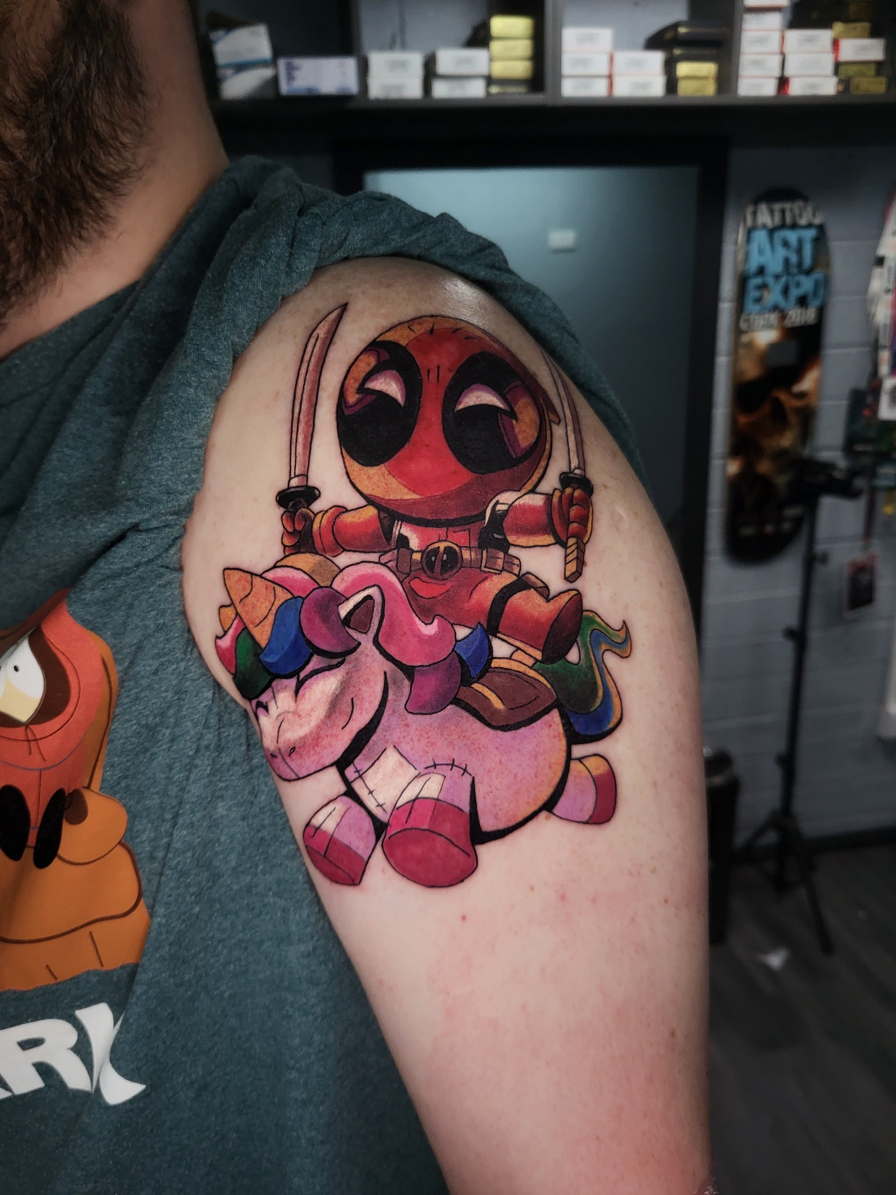 Kolorowy tatuaz Deadpoola w stylu cartoon na jednorożcu na ramieniu