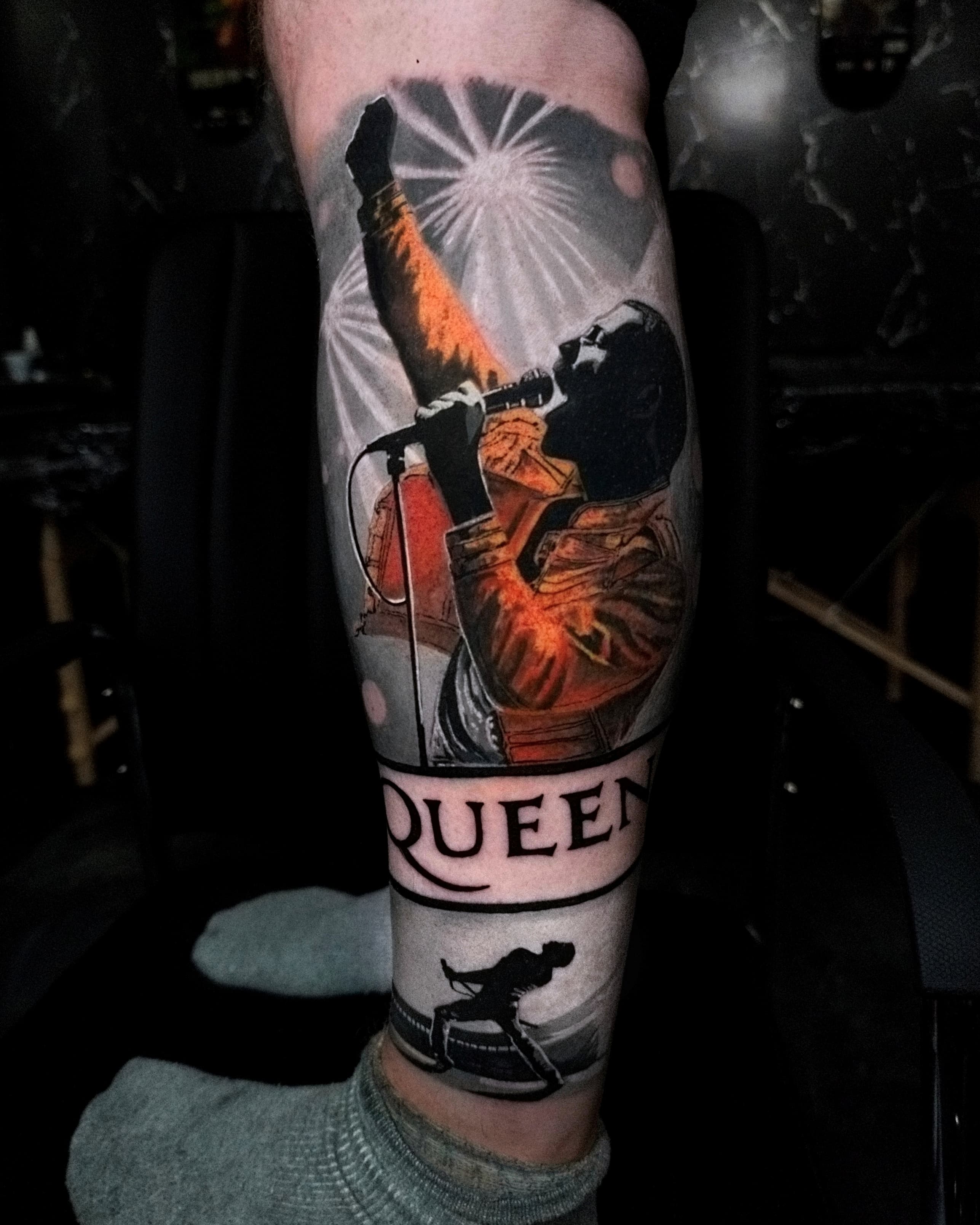 Kolorowy realistyczny tatuaż Freddiego Mercury'ego z logo Queen na przedramieniu z pomarańczowymi akcentami