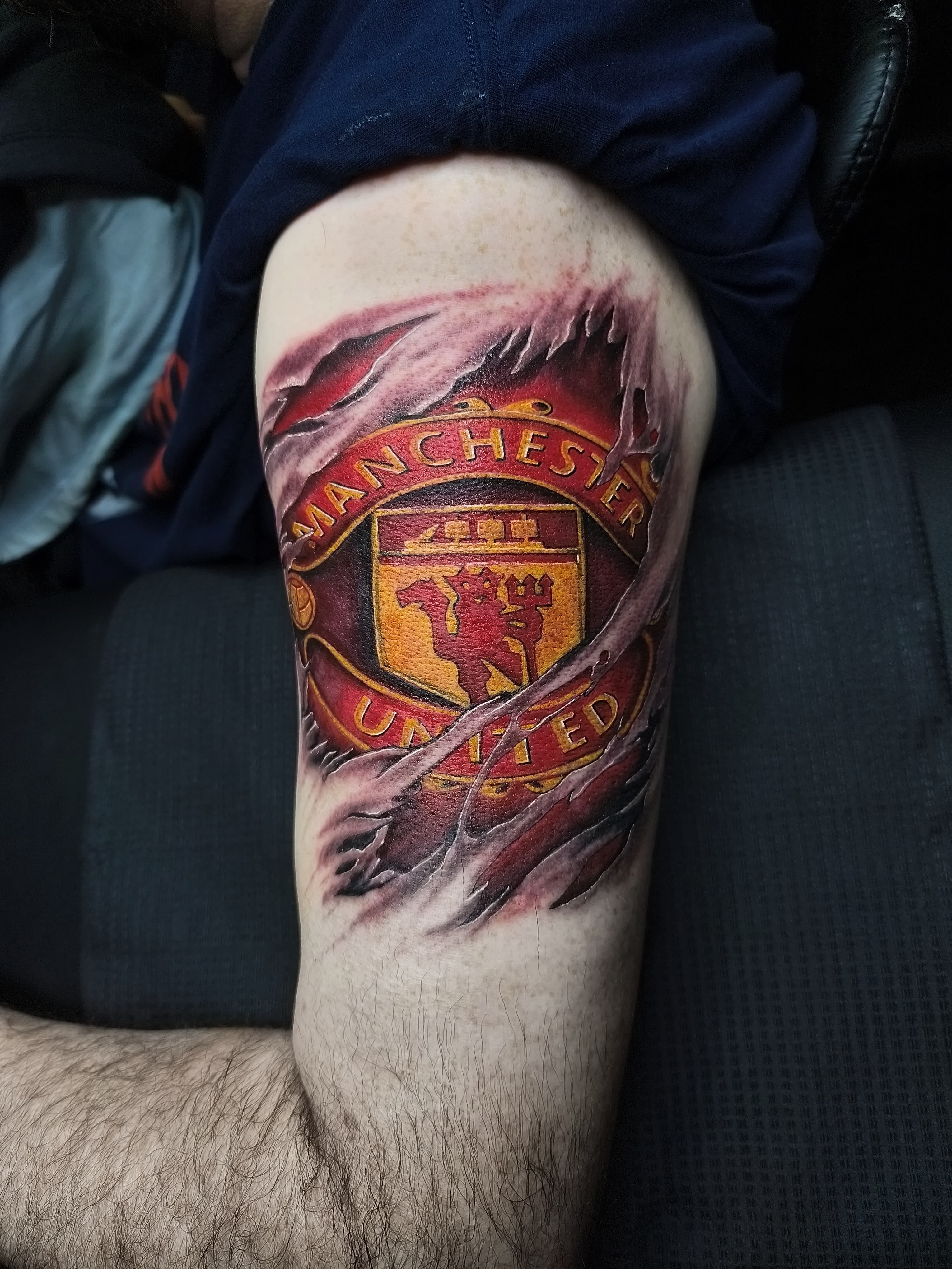 Kolorowy realistyczny tatuaz herbu Manchester United na udzie wyłaniający się spod rozdarej skóry