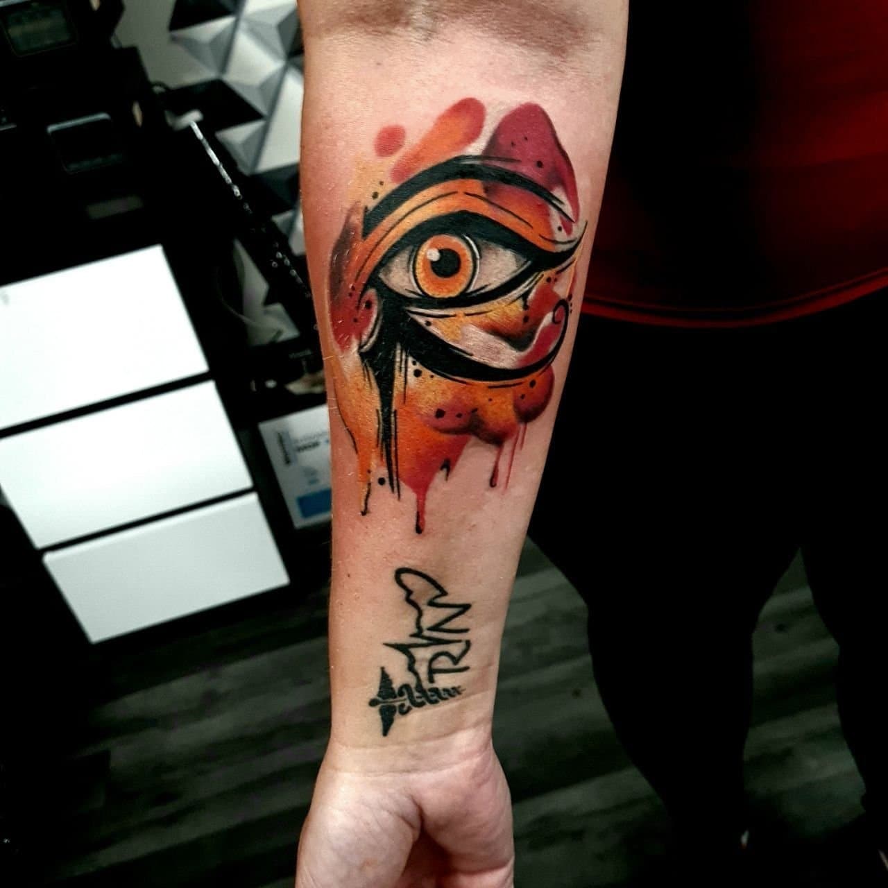 Kolorowy tatuaz oka Horusa w stylu watercolor z pomaranczowymi plamami na przedramieniu