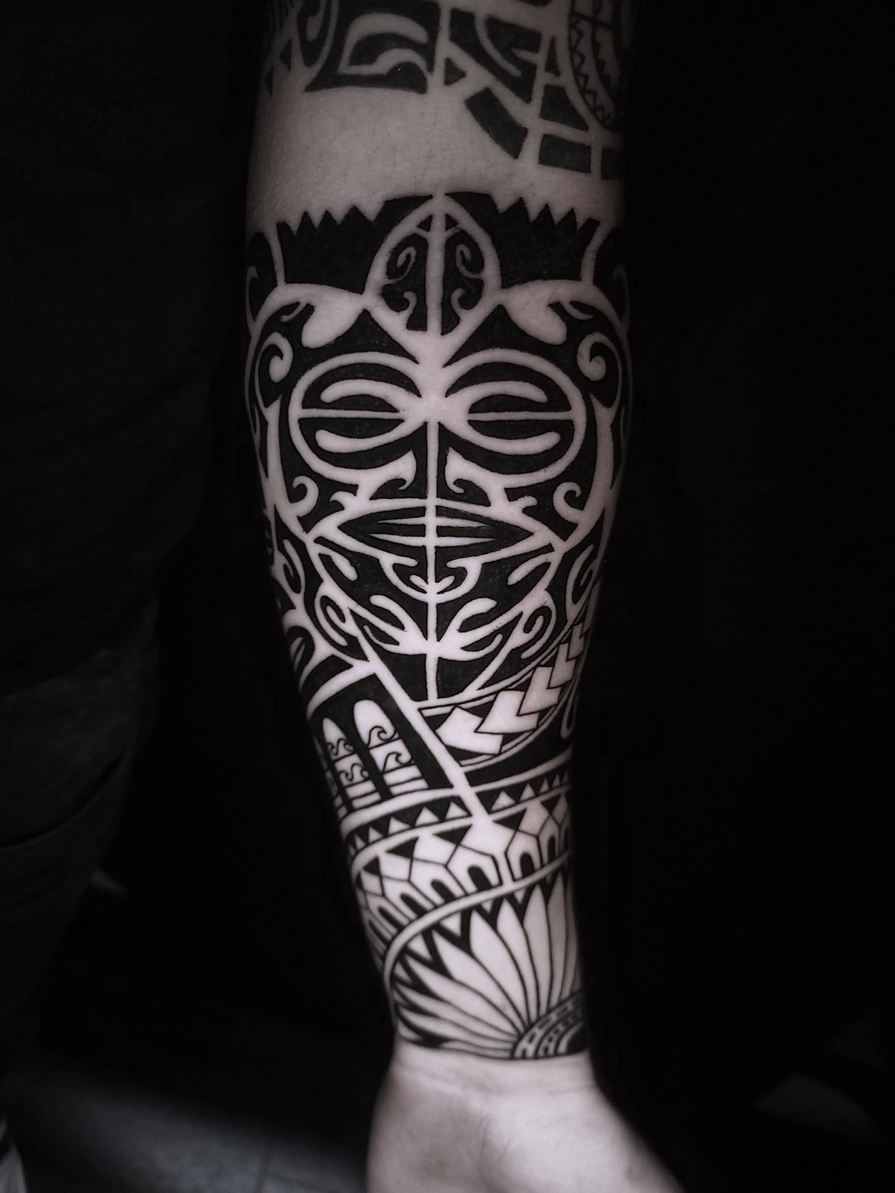 Czarny polinezyjski tatuaz z maska tiki i tradycyjnymi wzorami na przedramieniu
