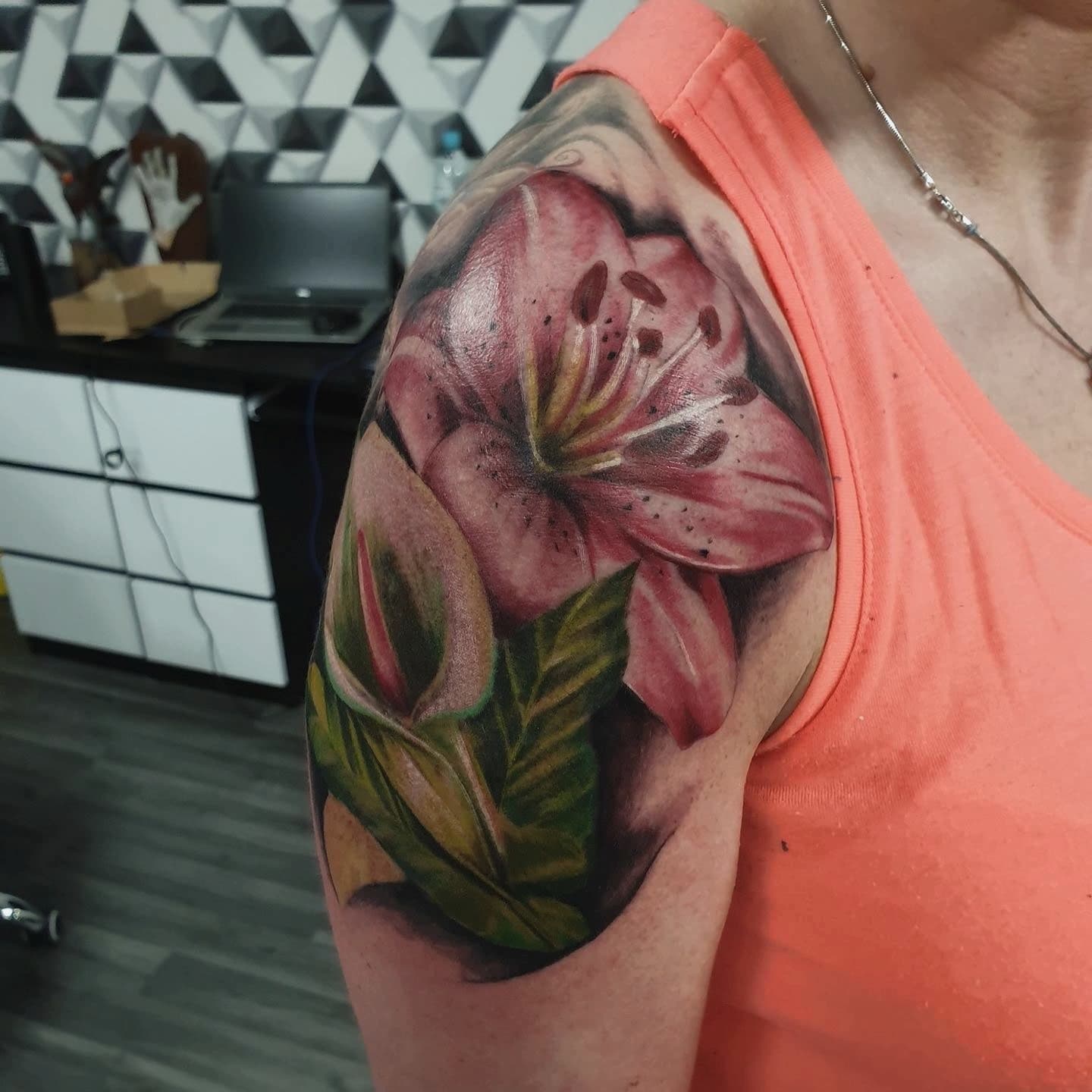 Kolorowy realistyczny tatuaz rozowej lilii z zielonymi liscmi na ramieniu kobiety