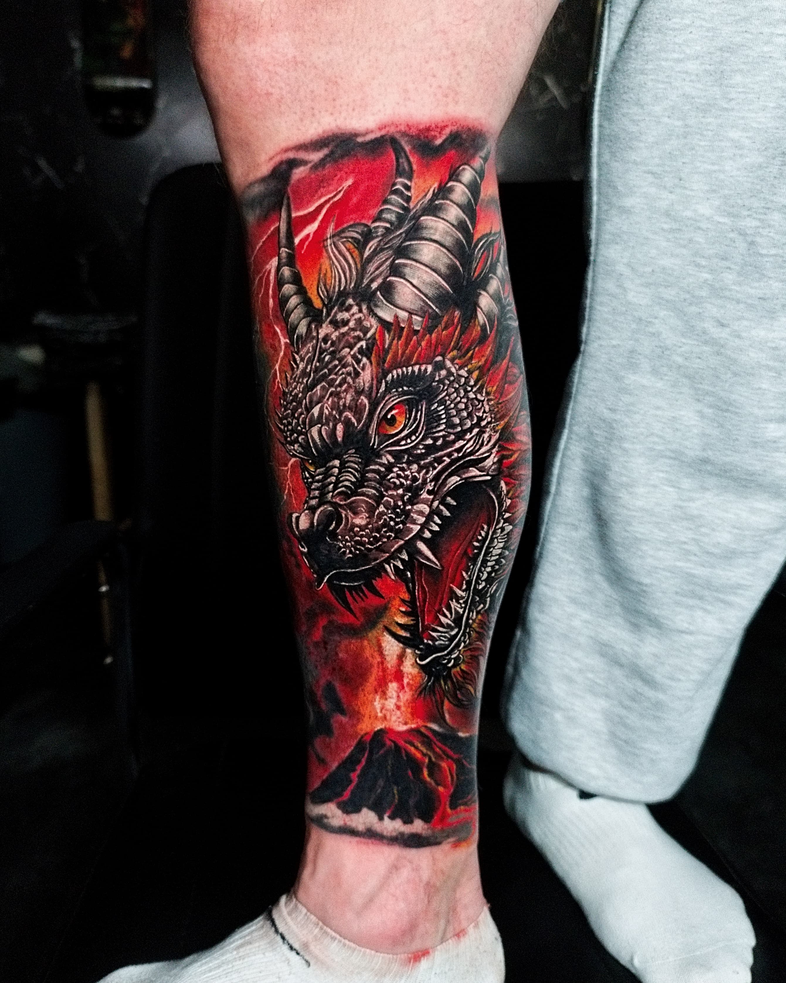Kolorowy realistyczny tatuaz głowy smoka z rogami na przedramieniu z ognistym czerwonym tłem